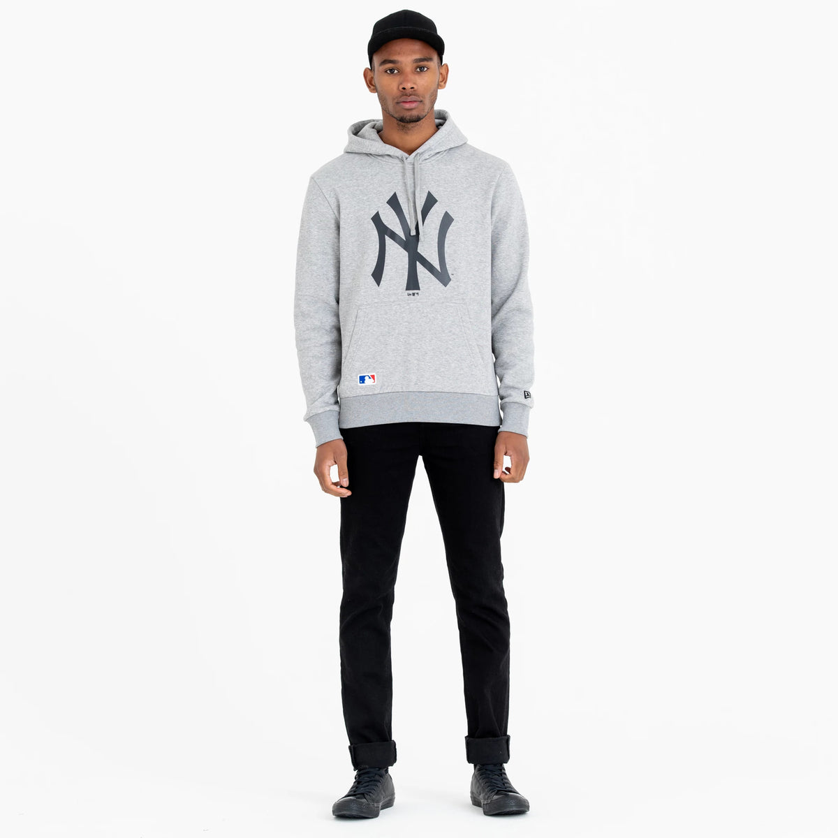 New York Yankees MLB Regular Grey Pullover Hoodie, on-model view showing the large black NY logo across the chest, a small raised logo patch near the cuff and a drawstring hood, worn with black trousers and shoes.