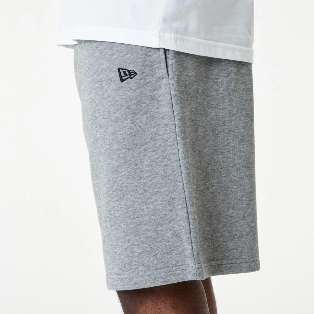 New Era Essential Grey Shorts, side view showing light grey fabric with a raised New Era logo in black near the pocket seam, the hem falling to knee length against a neutral backdrop.