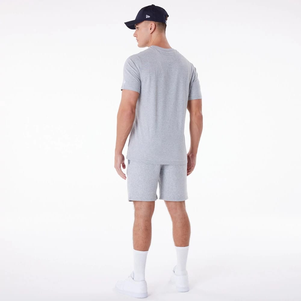 New Era Essential Grey Shorts, on-model rear view showing the light grey shorts paired with a matching grey T-shirt, white trainers, and socks, featuring a small debossed New Era logo on a navy cap worn by the model.