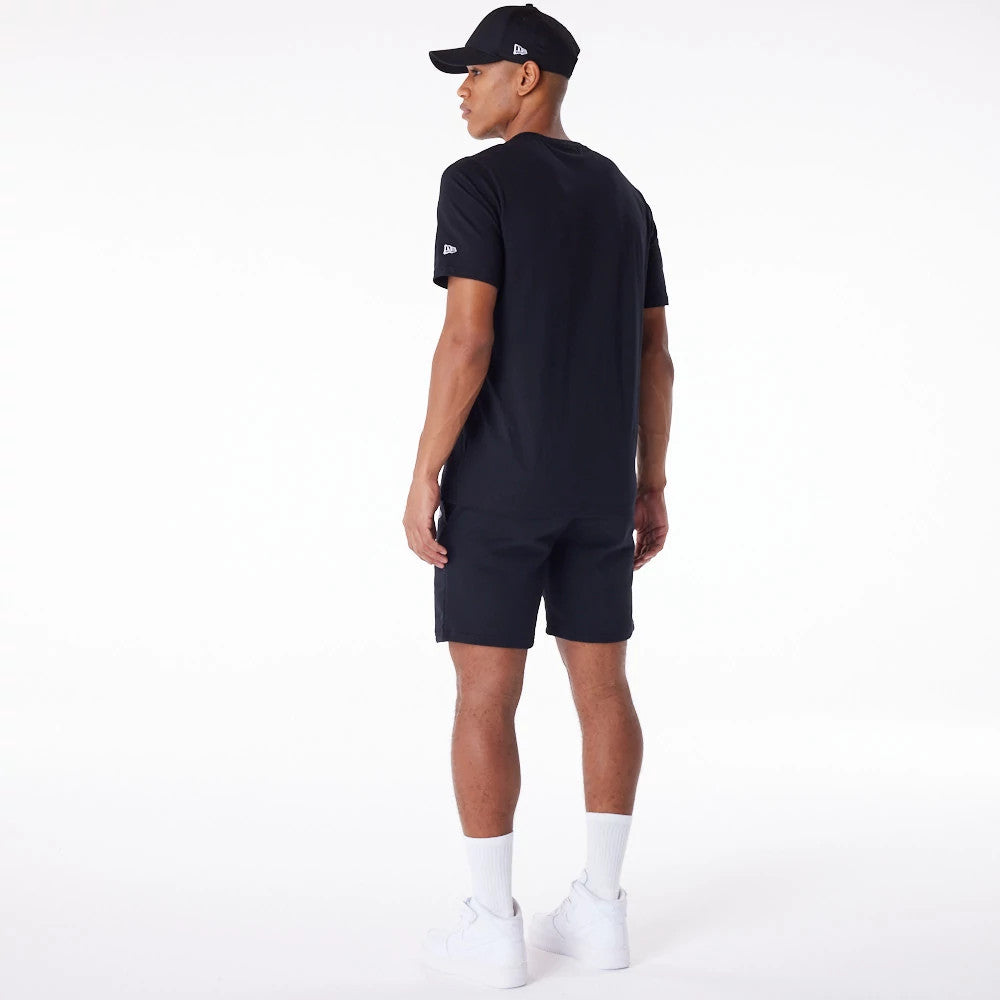 New Era Essential Black Logo Shorts, on-model view showing the black shorts worn with a matching black T-shirt and cap, a raised New Era logo on the left sleeve and side of the cap, paired with white socks and trainers.