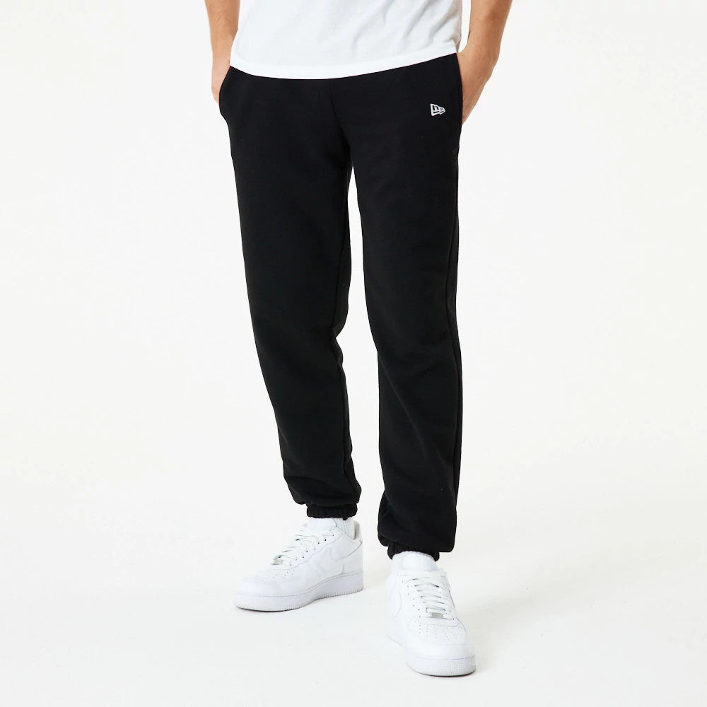 New Era Essential Black Joggers, on a model wearing a white T-shirt and white trainers, showing a relaxed fit with elasticated cuffs and the embroidered New Era flag logo on the left upper leg.