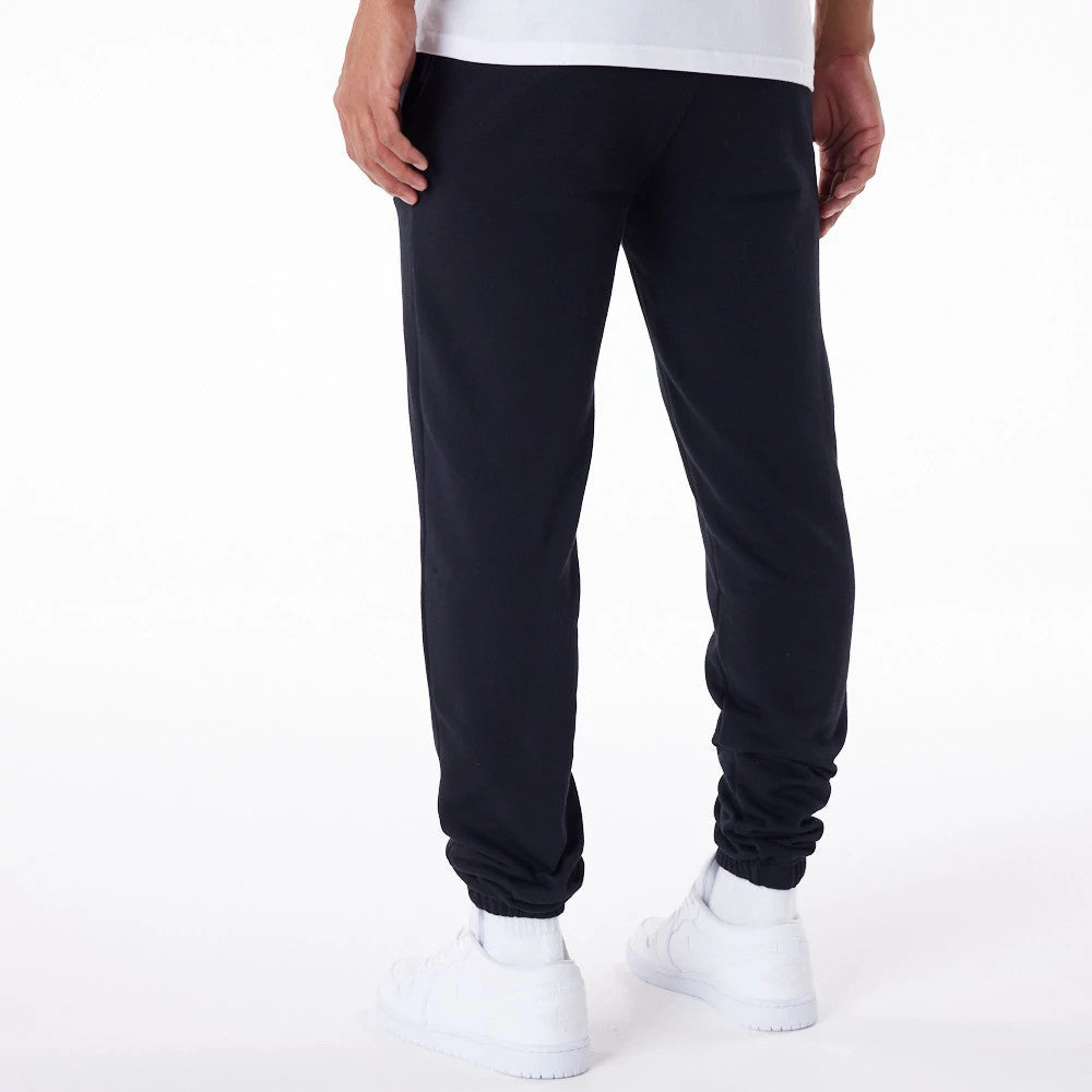 New Era Essential Black Joggers, back view on a model wearing white socks and shoes, highlighting an elasticated waistband, cuffed hems, and a relaxed fit extending from the hips to the ankles.