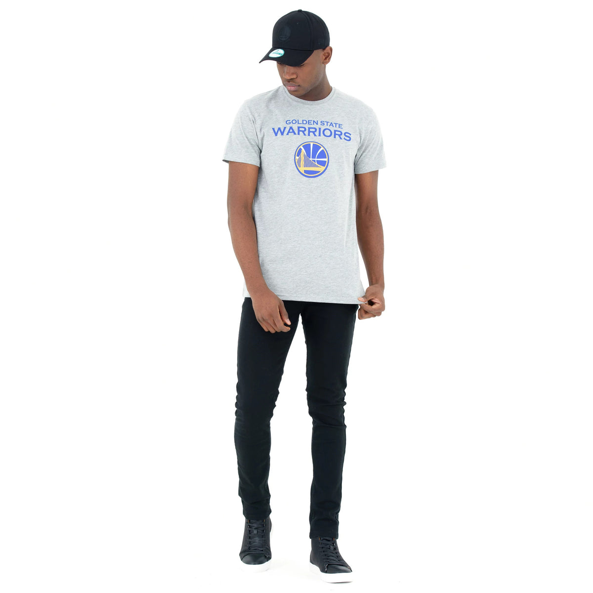 Golden State Warriors NBA Regular Grey T-Shirt, on-model view showing a grey T-shirt with blue "Golden State Warriors" text and a yellow basketball logo on the chest, worn with black jeans and a black cap with a debossed New Era logo.