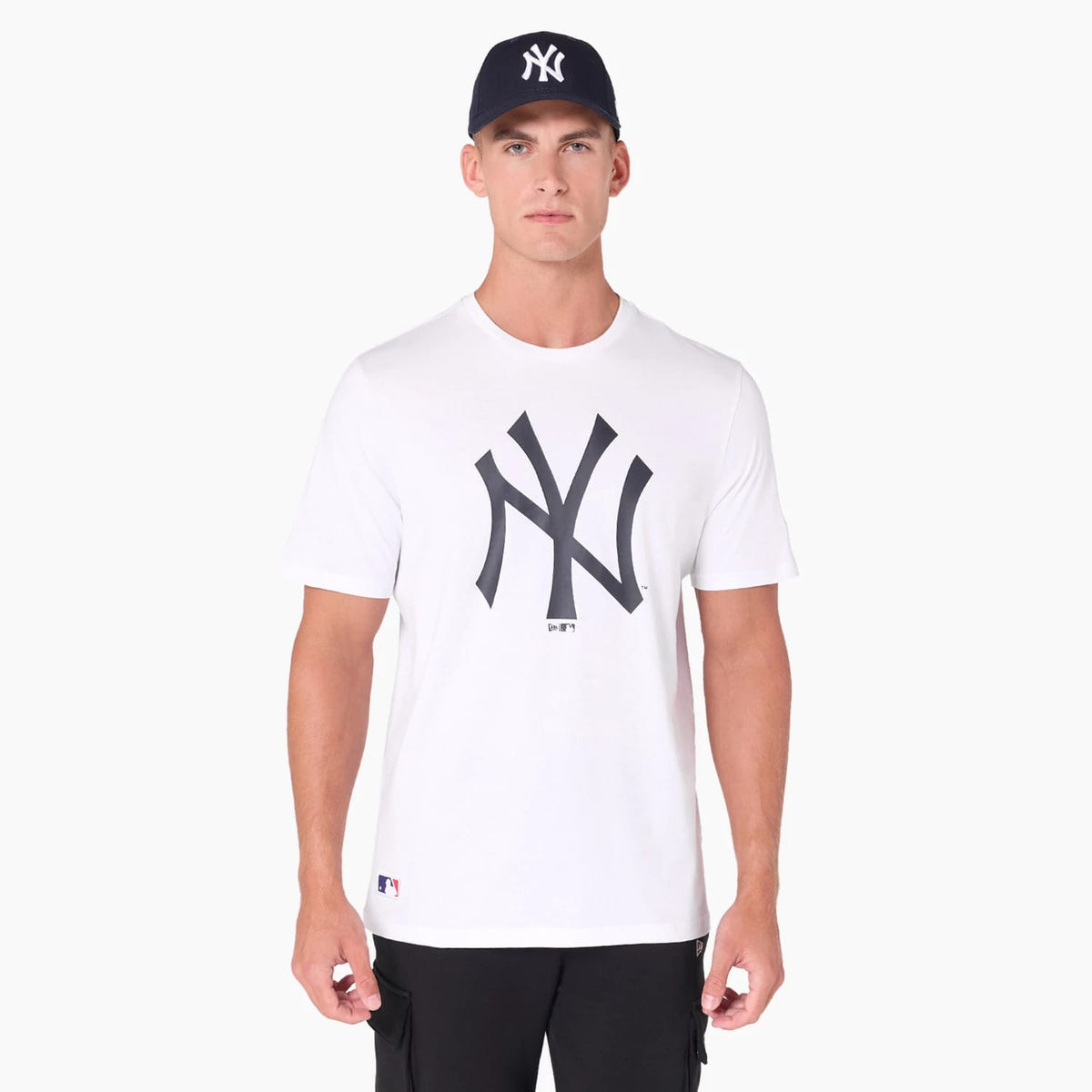 New York Yankees MLB White T-Shirt, front view showing a large black NY logo printed on the centre of the chest, a small MLB logo near the hem, and paired with a navy cap featuring a raised NY logo.