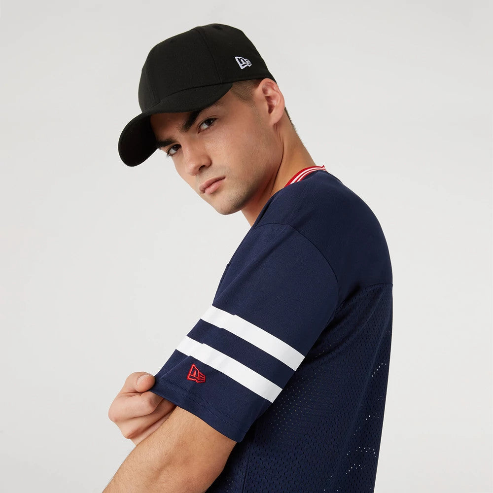 New England Patriots NFL Dark Blue Mesh T-Shirt, on-model view showing the dark blue mesh fabric with white stripe details on the sleeve, red embroidered New Era logo near the cuff, and contrasting collar trim, worn with a black cap featuring a raised logo on the side.