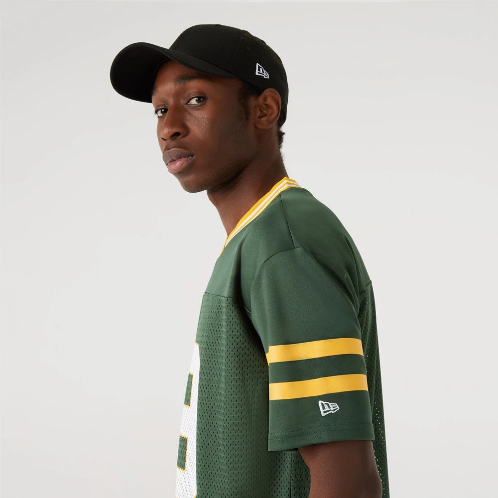 Green Bay Packers NFL Dark Green Mesh T-Shirt, on-model view showing the dark green shirt with yellow stripes on the sleeve, a white embroidered New Era logo above the cuff, and a black cap with a white raised logo on the side.