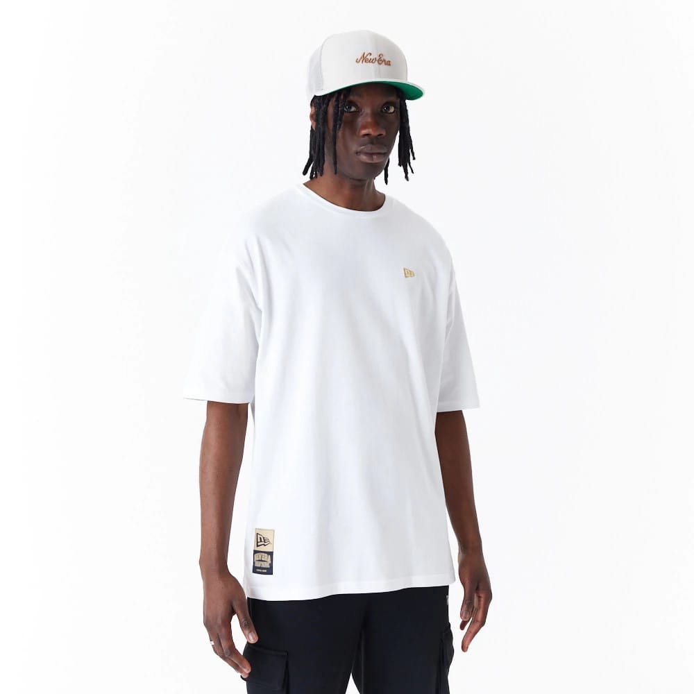 New Era Sticker White Oversized T-Shirt, on-model view showing the relaxed white fit with a small debossed gold New Era logo on the left chest and a larger patch near the bottom hem.