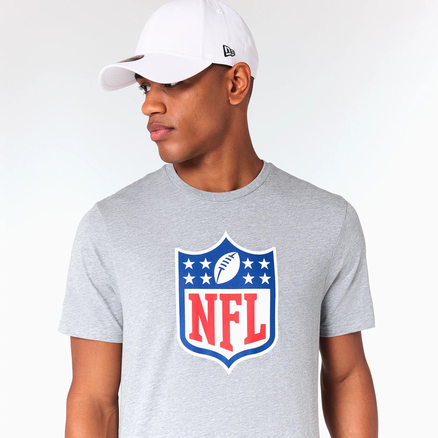New Era NFL Shield Logo NFL Grey T-Shirt, on-model view showing the large red and blue NFL shield emblem centred on the chest, worn with a plain white cap featuring a raised New Era logo on the side.