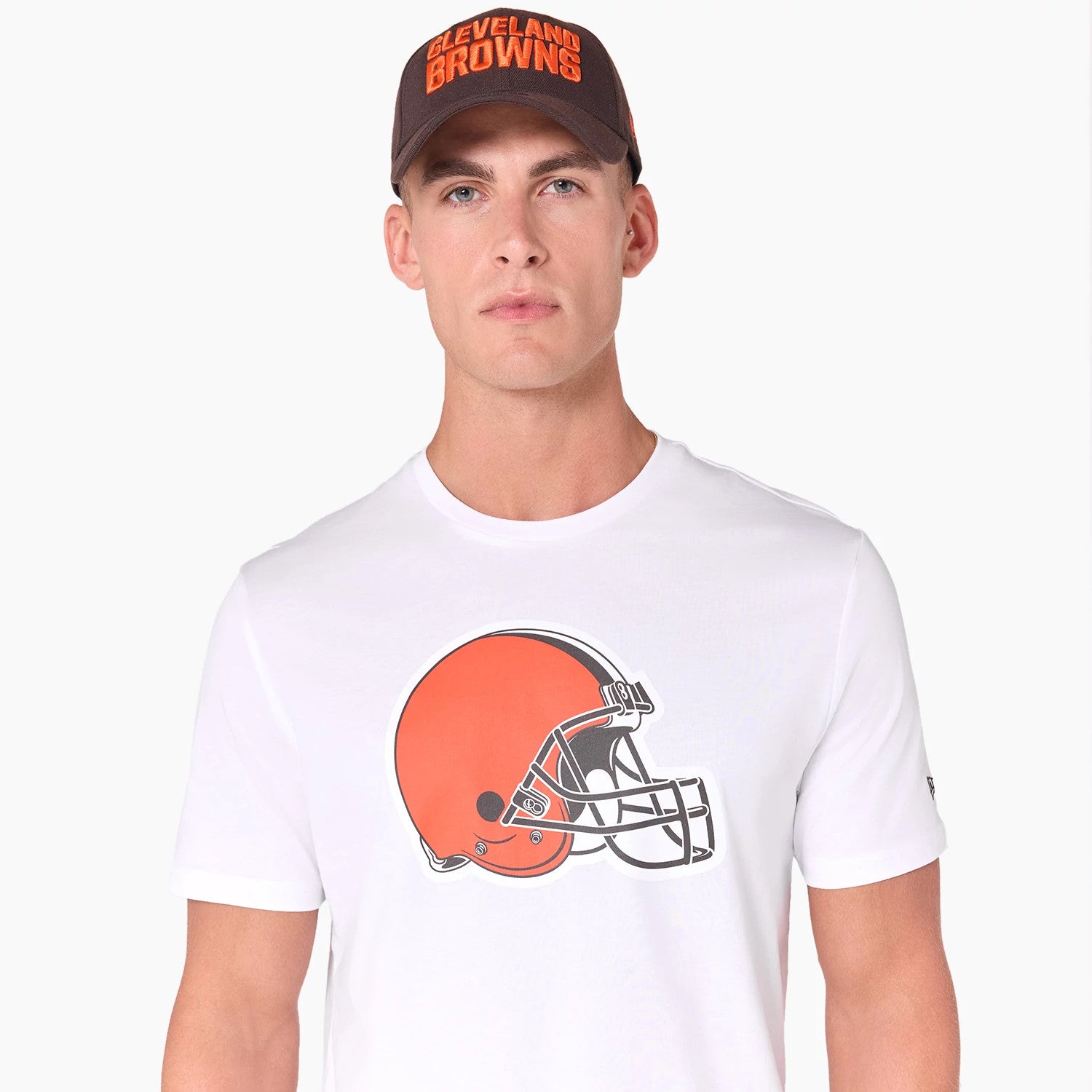 New Era Cleveland Browns NFL White T-Shirt, on-model view showing a large orange helmet graphic centred on the chest, a black embroidered New Era logo on the left sleeve and a brown cap with Cleveland Browns text worn on the head.