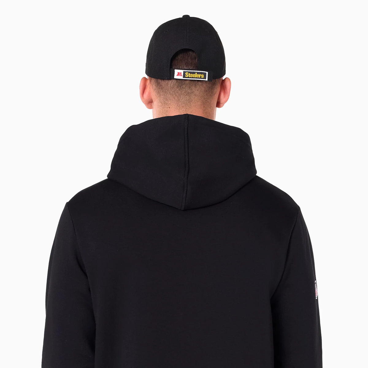 New Era Pittsburgh Steelers NFL Black Pullover Hoodie, rear view showing the plain black hood and shoulders, worn by a person with a black cap featuring a raised Steelers logo patch above the adjustable strap opening at the back.