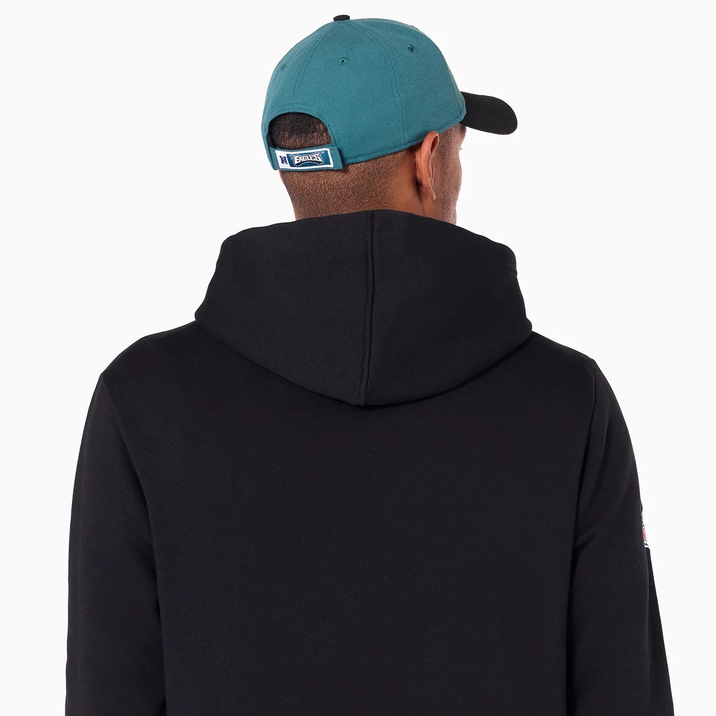 New Era Philadelphia Eagles NFL Black Pullover Hoodie, rear view showing the black hood resting against the back and a person wearing a turquoise cap with an embroidered Philadelphia Eagles logo tag above the strap.