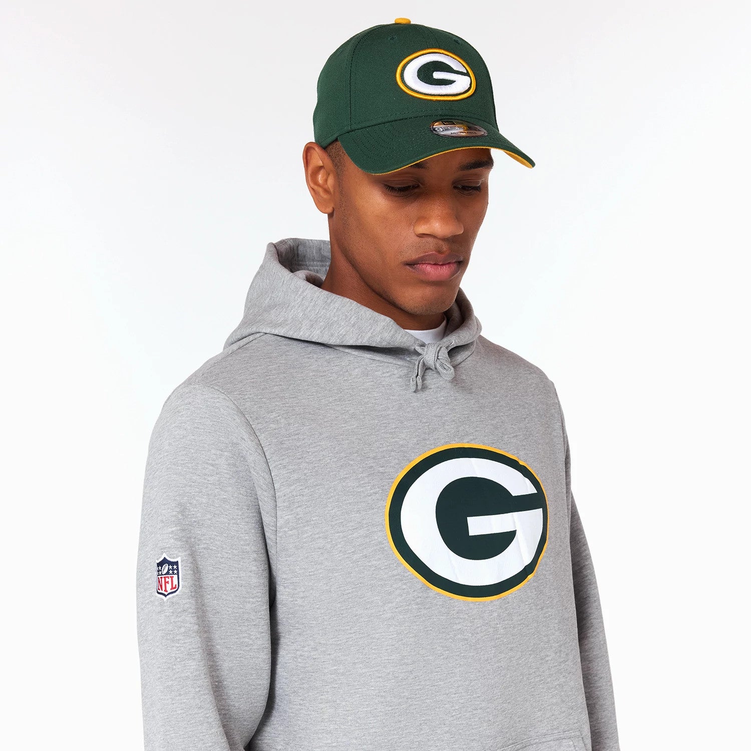 New Era Green Bay Packers NFL Grey Pullover Hoodie, on-model view showing a large printed Packers G logo in green and white on the chest, an NFL logo patch on the sleeve, and a matching green cap worn on the head.