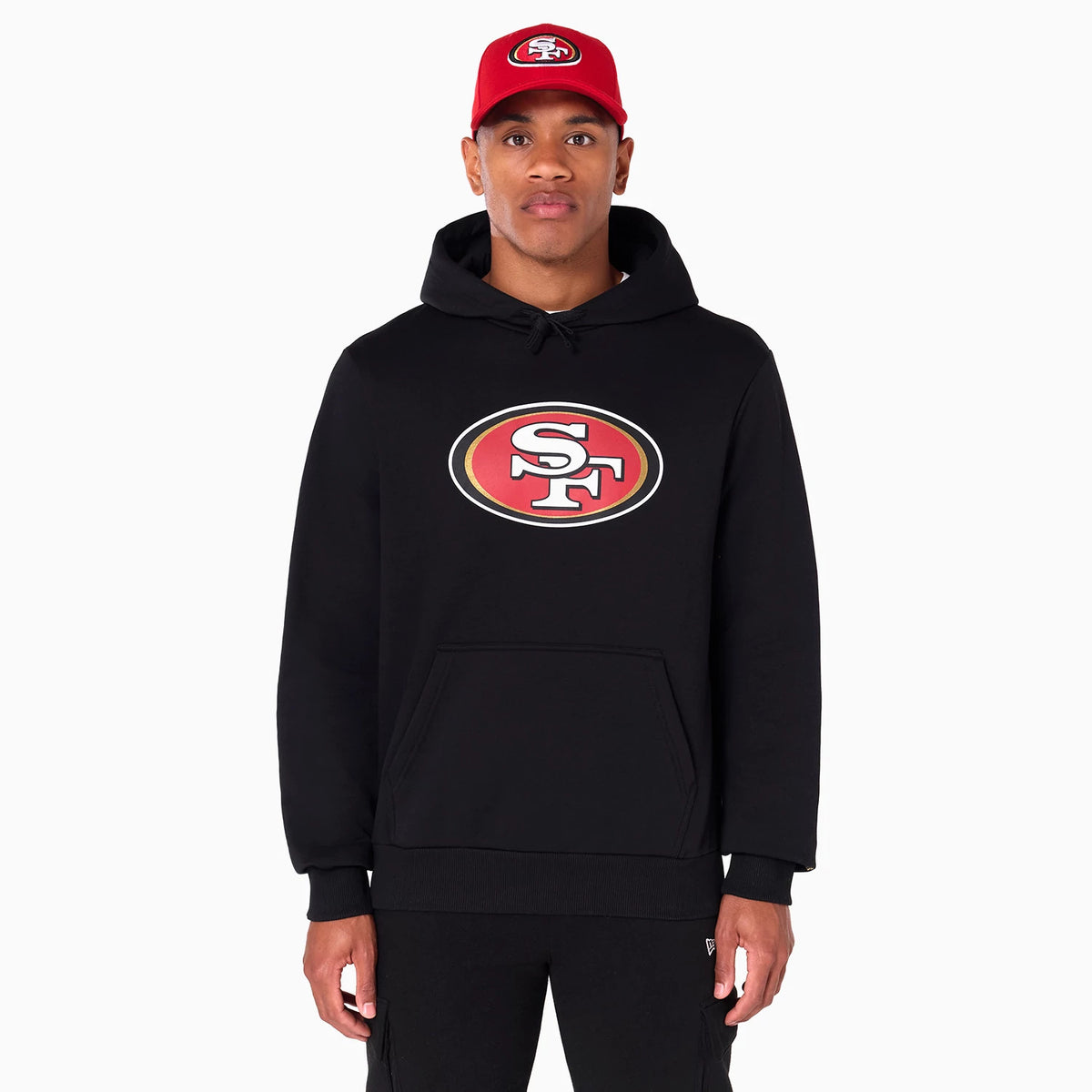 New Era San Francisco 49ers NFL Black Pullover Hoodie, on-model view showing the large SF logo centred on the chest of the black hoodie, the hood resting on the shoulders and eyelets around the neckline, worn with a red cap featuring a raised logo.