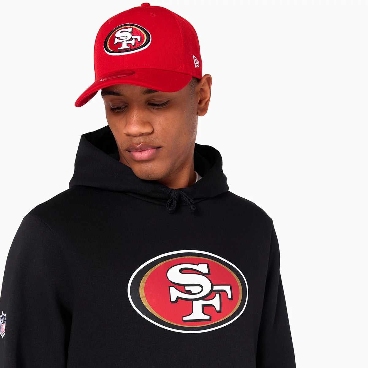 New Era San Francisco 49ers NFL Black Pullover Hoodie, on-model view showing a large printed team logo on the chest of the black hoodie, worn under a red cap with an embossed SF logo on the front panel.