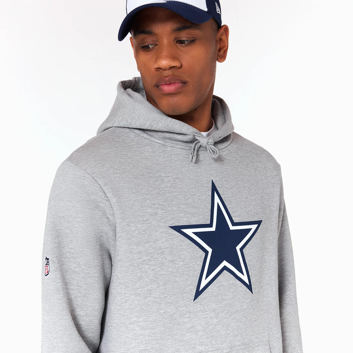 New Era Dallas Cowboys NFL Grey Pullover Hoodie, on-model view showing a dark blue star emblem centred on the chest, drawstrings on the hood, an NFL patch embroidered on the left sleeve, and the model wearing a navy and white cap.