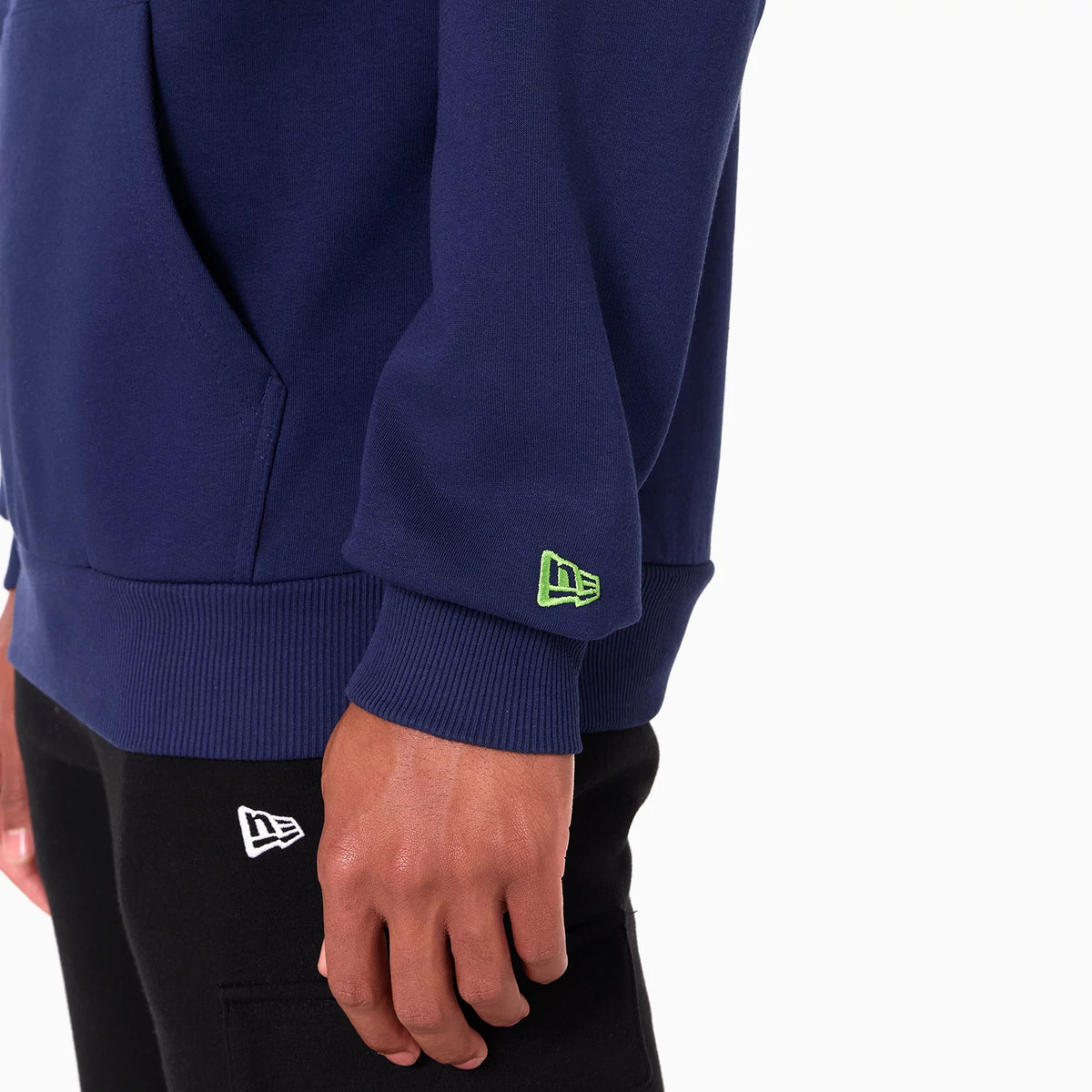 New Era Seattle Seahawks NFL Navy Pullover Hoodie, on-model view showing the navy sleeve with a green embroidered New Era logo near the cuff and a front pouch pocket, worn with black trousers featuring a white embroidered New Era logo.