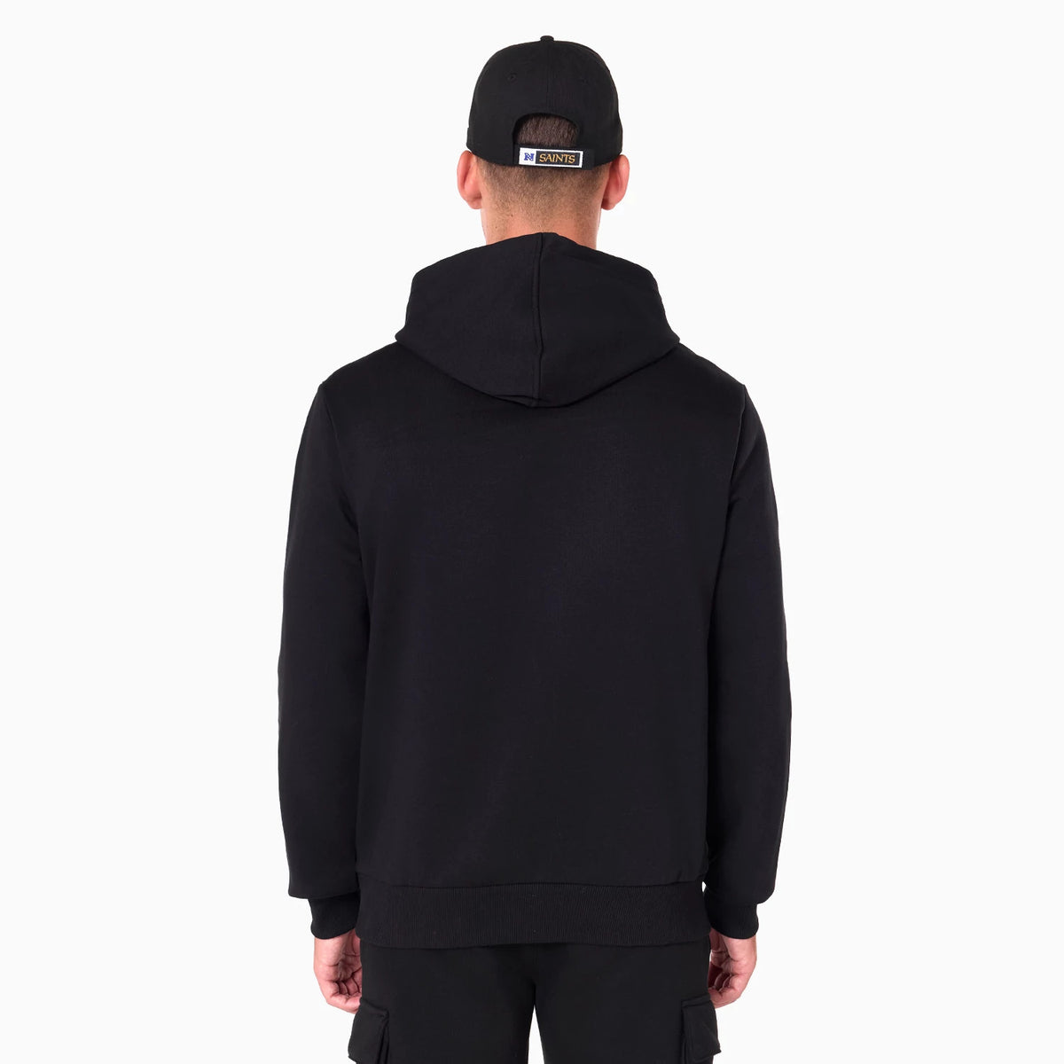 New Era New Orleans Saints NFL Black Pullover Hoodie, rear view showing the plain black back of the hoodie with the hood down, worn by a male model in black trousers, a black cap with a raised patch on the back.