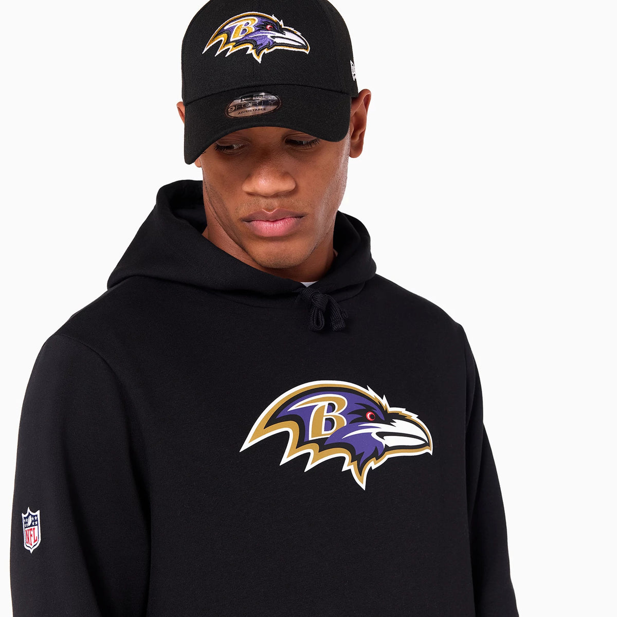 New Era Baltimore Ravens NFL Black Pullover Hoodie, on-model view showing the large Baltimore Ravens logo printed on the chest, a small NFL logo patch on the sleeve, and a matching cap with a similar raised logo on the front.