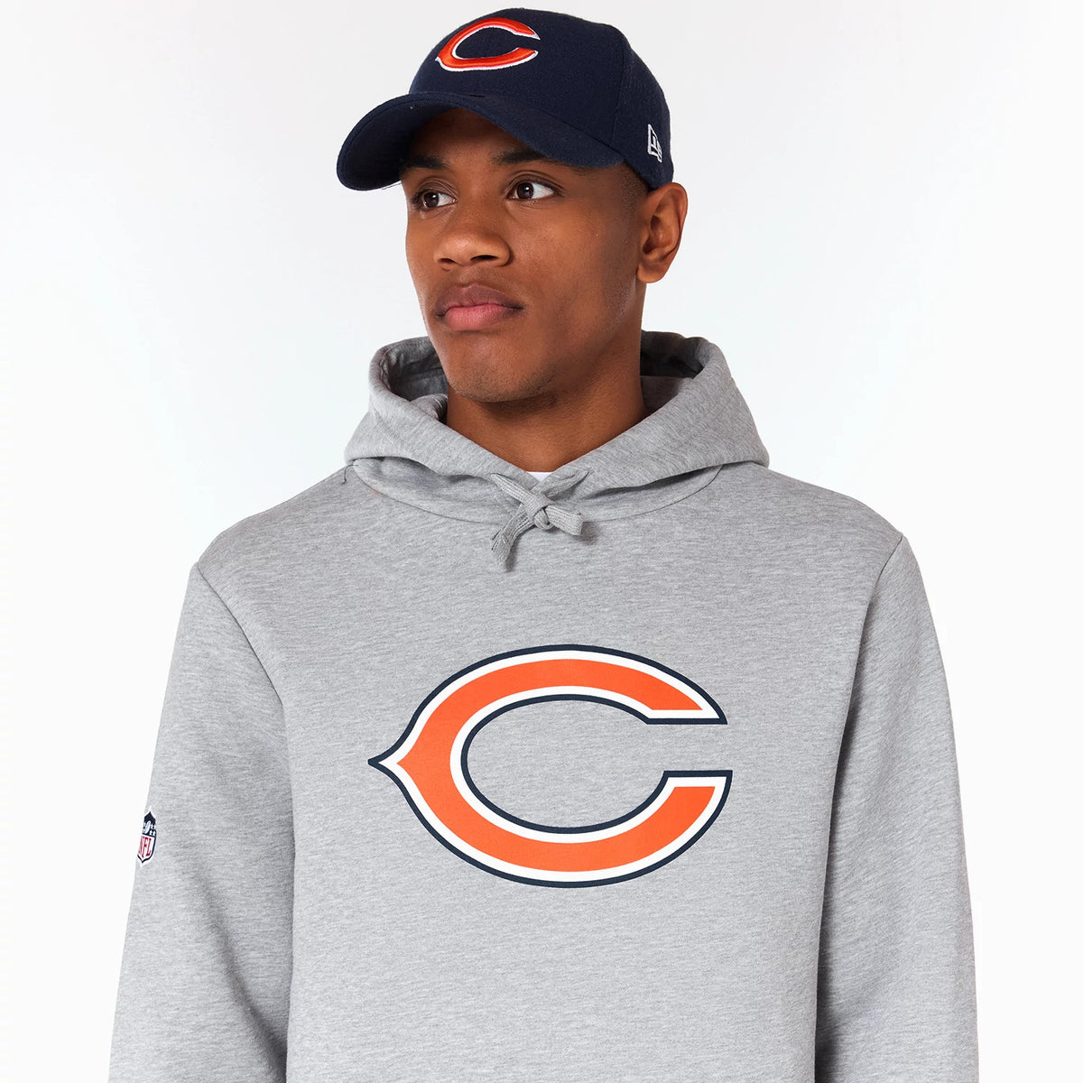 New Era Chicago Bears NFL Grey Pullover Hoodie, on-model view showing the large orange and navy team logo centred on the chest, with a small NFL logo on the left sleeve, paired with a navy cap featuring an orange raised logo.