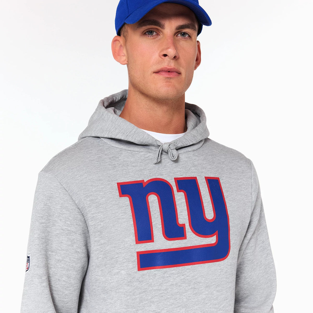 New Era New York Giants NFL Grey Pullover Hoodie, front view showing a large blue and red 'ny' logo on the chest, a drawstring hood, and an NFL patch on the left sleeve, worn with a blue cap.