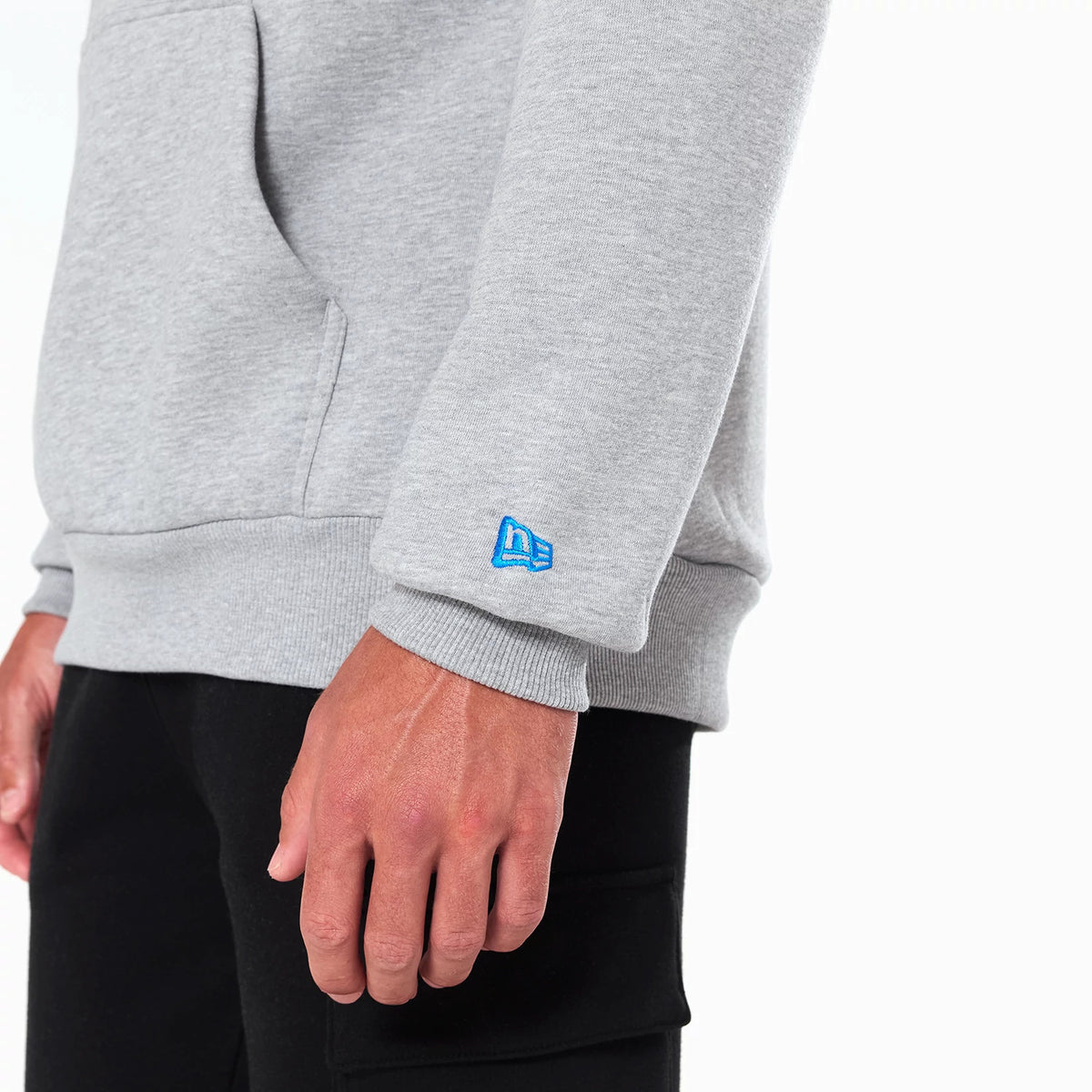 New Era Detroit Lions NFL Grey Pullover Hoodie, side view showing the light grey fabric with a raised blue New Era logo on the cuff, a pocket seam along the side, and ribbed cuffs and hem against black trousers.
