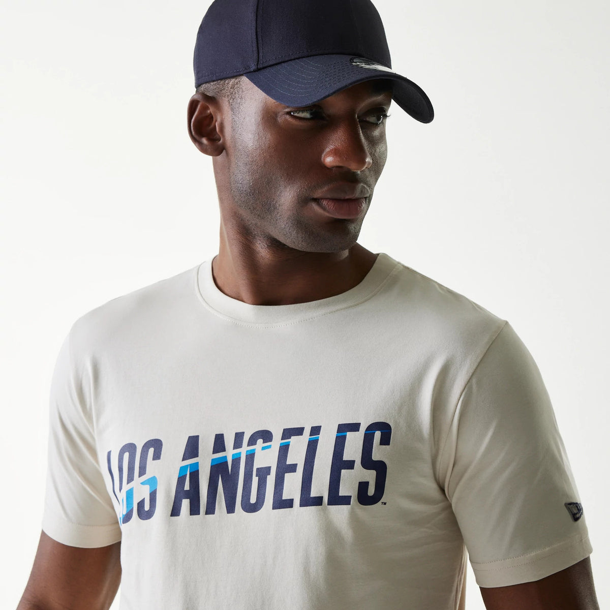 New Era LA Dodgers MLB City Connect Cream T-Shirt, on-model view showing the cream fabric with bold blue Los Angeles text across the chest and a small New Era logo on the left sleeve, paired with a navy cap.
