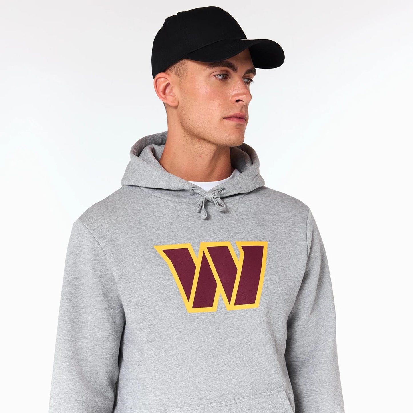 New Era Washington Commanders NFL Grey Pullover Hoodie, on-model view showing the grey hoodie with a large yellow and maroon W logo printed on the chest, drawstrings hanging at the neck, and a black cap worn by the model.