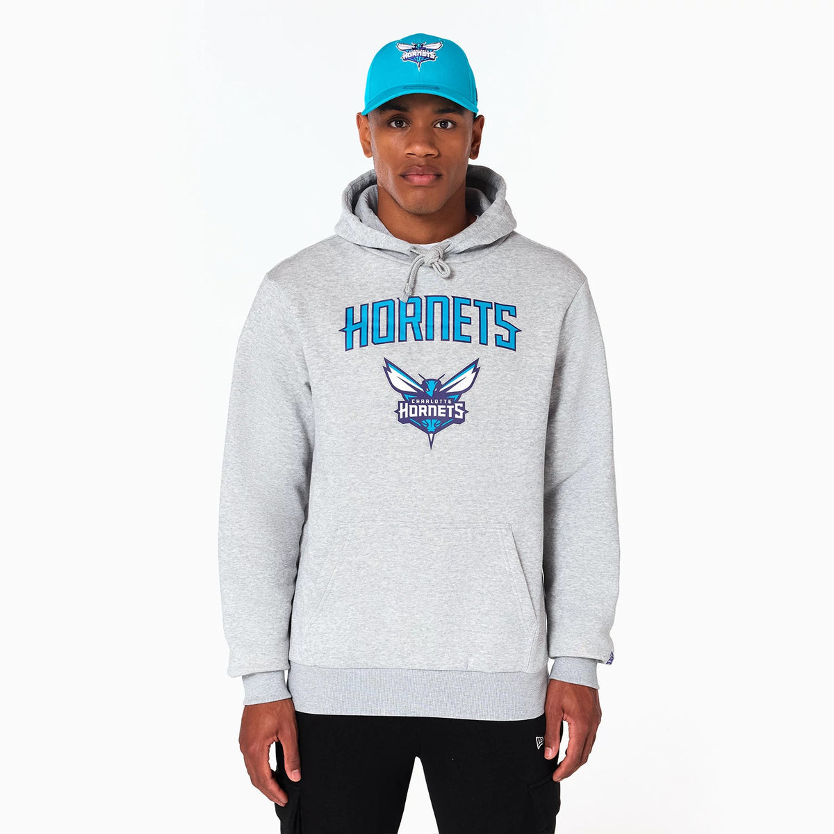 New Era Charlotte Hornets NBA Grey Pullover Hoodie, on-model view showing the front in light grey with HORNETS in blue across the chest, a large team logo below, and a small debossed New Era logo on the left sleeve cuff.