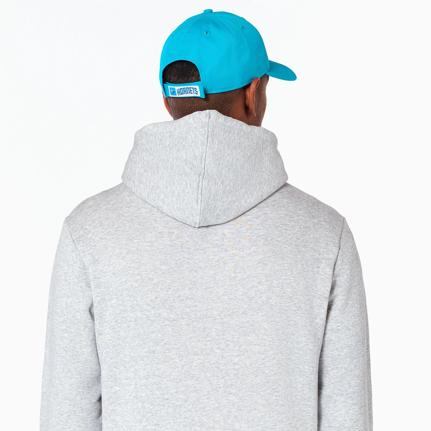 New Era Charlotte Hornets NBA Grey Pullover Hoodie, rear view showing the light grey hood against the back, worn by a person in a blue cap with an embossed Hornets logo on the adjustable strap.