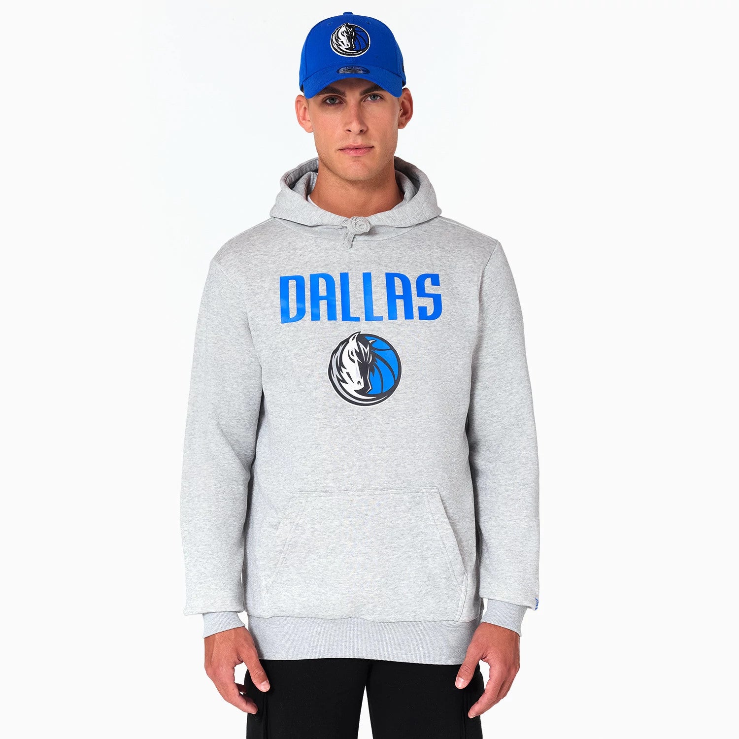 New Era Dallas Mavericks NBA Grey Pullover Hoodie, on-model view showing the grey hoodie with DALLAS in blue across the chest above the Mavericks logo, paired with a blue cap featuring a raised Mavericks logo on the front.