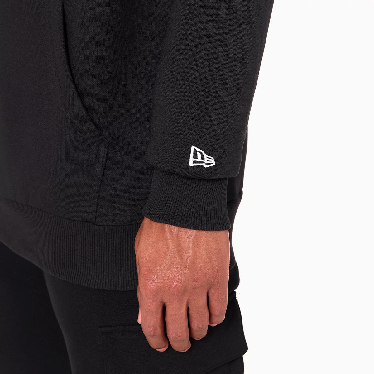 New Era San Antonio Spurs NBA Black Pullover Hoodie, side view showing the black sleeve with a white embroidered New Era logo near the cuff, and part of the hand in black trousers.