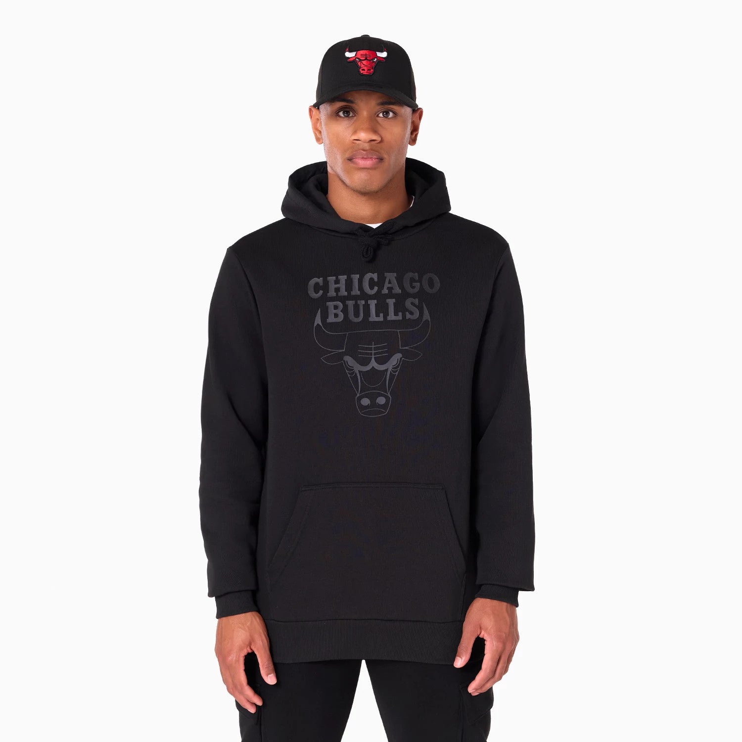 New Era Chicago Bulls NBA Black Pullover Hoodie, on-model view showing the black hoodie with a large front pocket, featuring a grey Chicago Bulls graphic centred on the chest, worn by a person also wearing a black cap with a red raised Bulls logo.