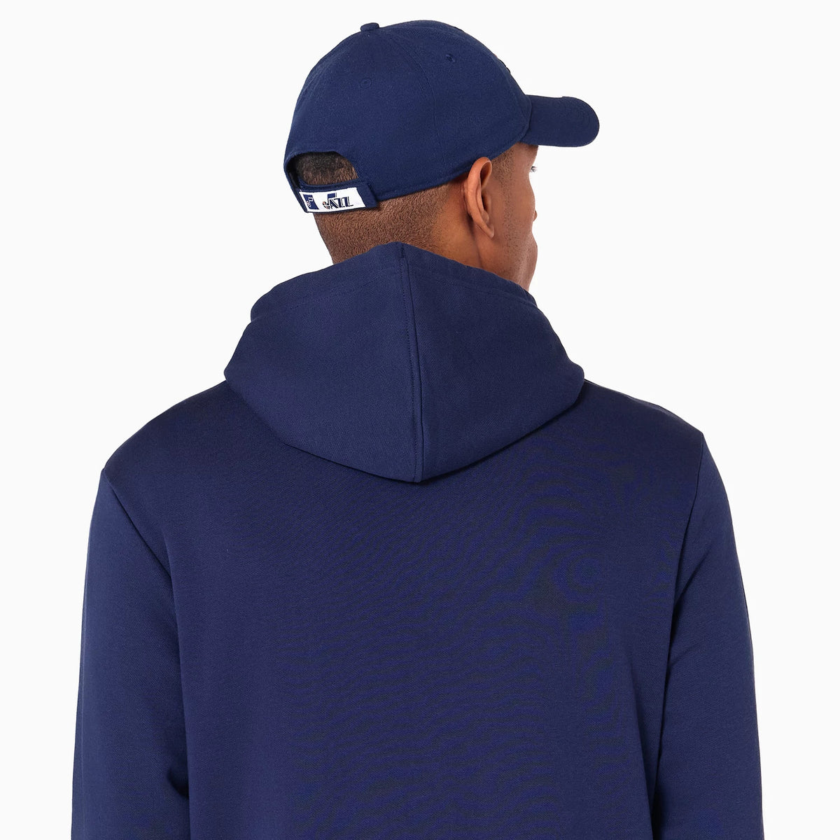 New Era Utah Jazz NBA Navy Pullover Hoodie, on-model rear view showing the hooded navy garment from the back paired with a navy cap featuring a raised logo above the adjustable strap, and rows of eyelets on top of the cap.