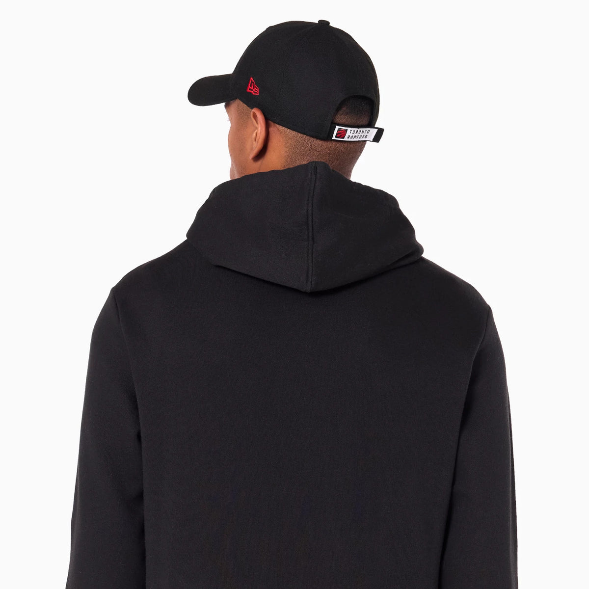 New Era Toronto Raptors NBA Black Pullover Hoodie, rear view showing a man wearing the hoodie with the hood down, paired with a black cap that has a red debossed New Era logo on the side and a Toronto Raptors tag at the back.