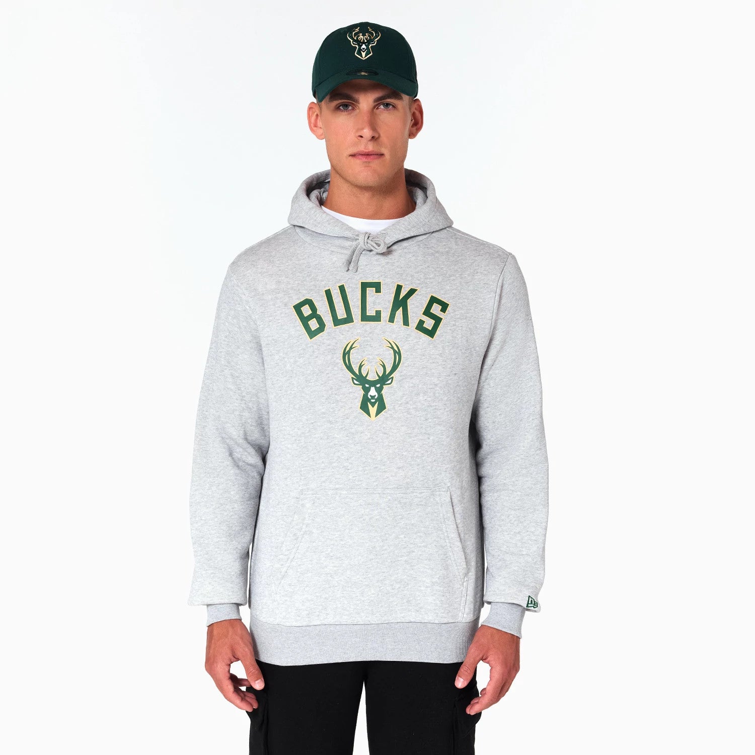 New Era Milwaukee Bucks NBA Grey Pullover Hoodie, front view showing a light grey hoodie with BUCKS lettering and a deer logo in green on the chest, worn by a model also wearing a dark green cap featuring an embossed deer logo.