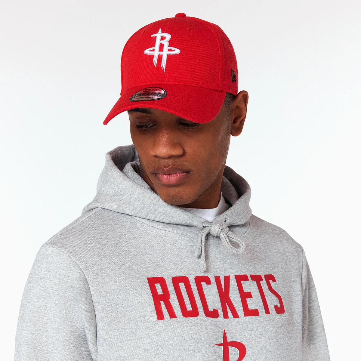 New Era Houston Rockets NBA Grey Pullover Hoodie, on-model view showing a grey hoodie with a red ROCKETS print across the chest, paired with a red cap featuring a raised white logo and multiple eyelets on each panel.