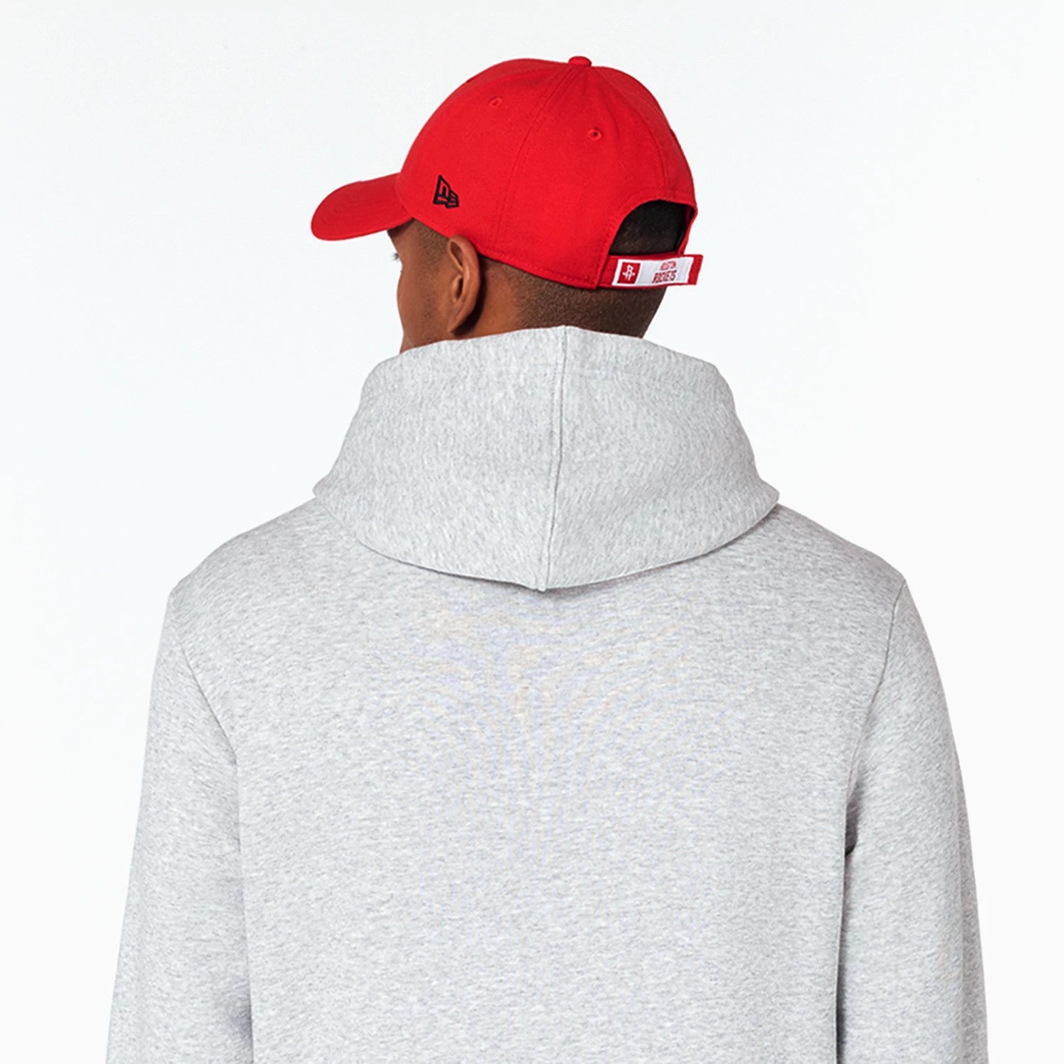 New Era Houston Rockets NBA Grey Pullover Hoodie, rear view showing the light grey fabric and large hood, worn by a model also wearing a red cap with a raised New Era logo on the side and adjustable strap at the back.