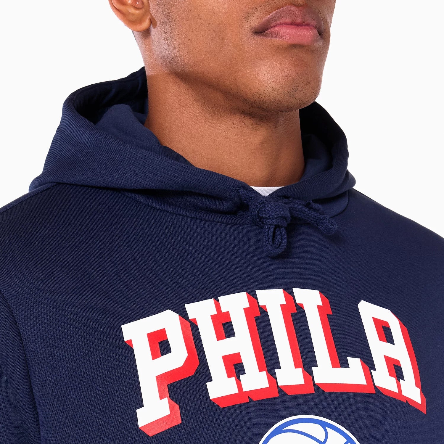 New Era Philadelphia 76ers NBA Navy Pullover Hoodie, on-model view showing the navy hooded top with a drawstring neckline, bold white and red letters spelling PHILA across the chest above a basketball graphic.
