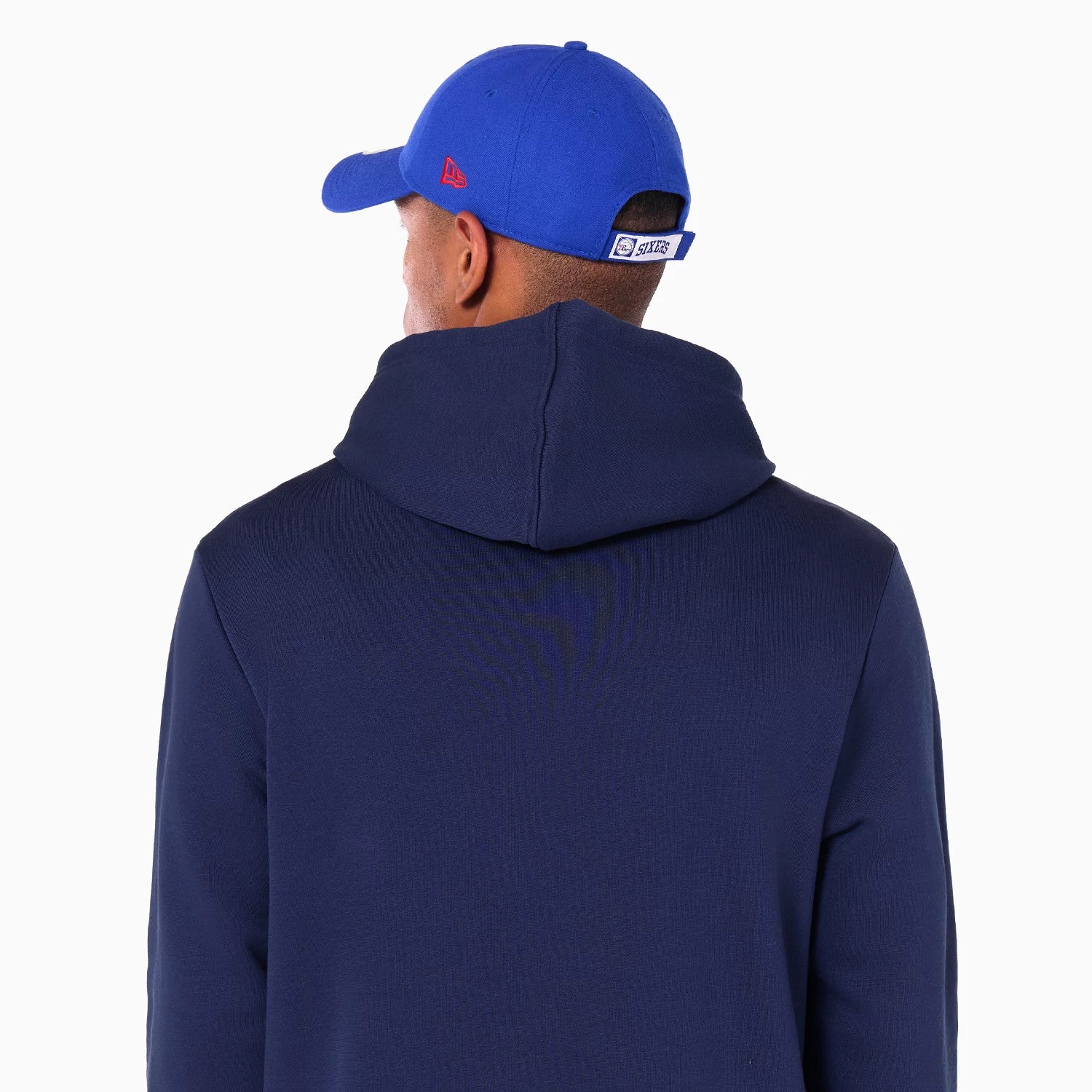 New Era Philadelphia 76ers NBA Navy Pullover Hoodie, rear view showing the dark navy hood and back, a bright blue cap above with a debossed New Era logo on the side panel in red and a sewn-on team patch on the adjustable strap.