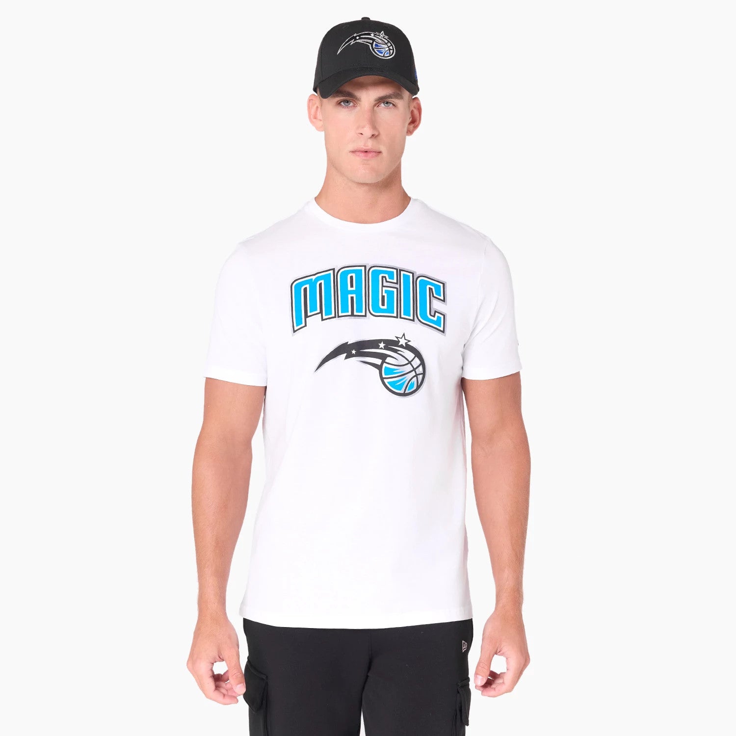 New Era Orlando Magic NBA White T-Shirt, front view showing large MAGIC text and team logo in blue and black on the chest, worn by a model paired with a black cap featuring a debossed Orlando Magic logo on the front.