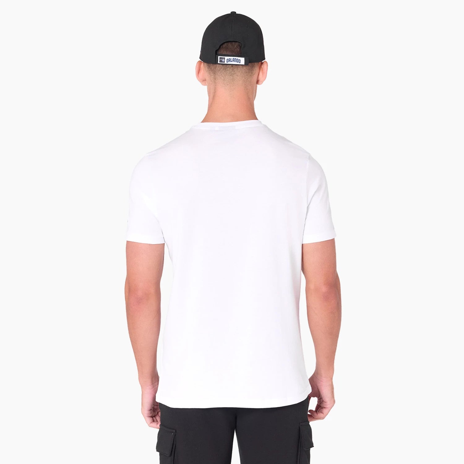 New Era Orlando Magic NBA White T-Shirt, on-model view showing the plain white shirt from the back, short sleeves and round neckline, paired with black cargo shorts and a black cap with an embossed logo on a rectangular patch.