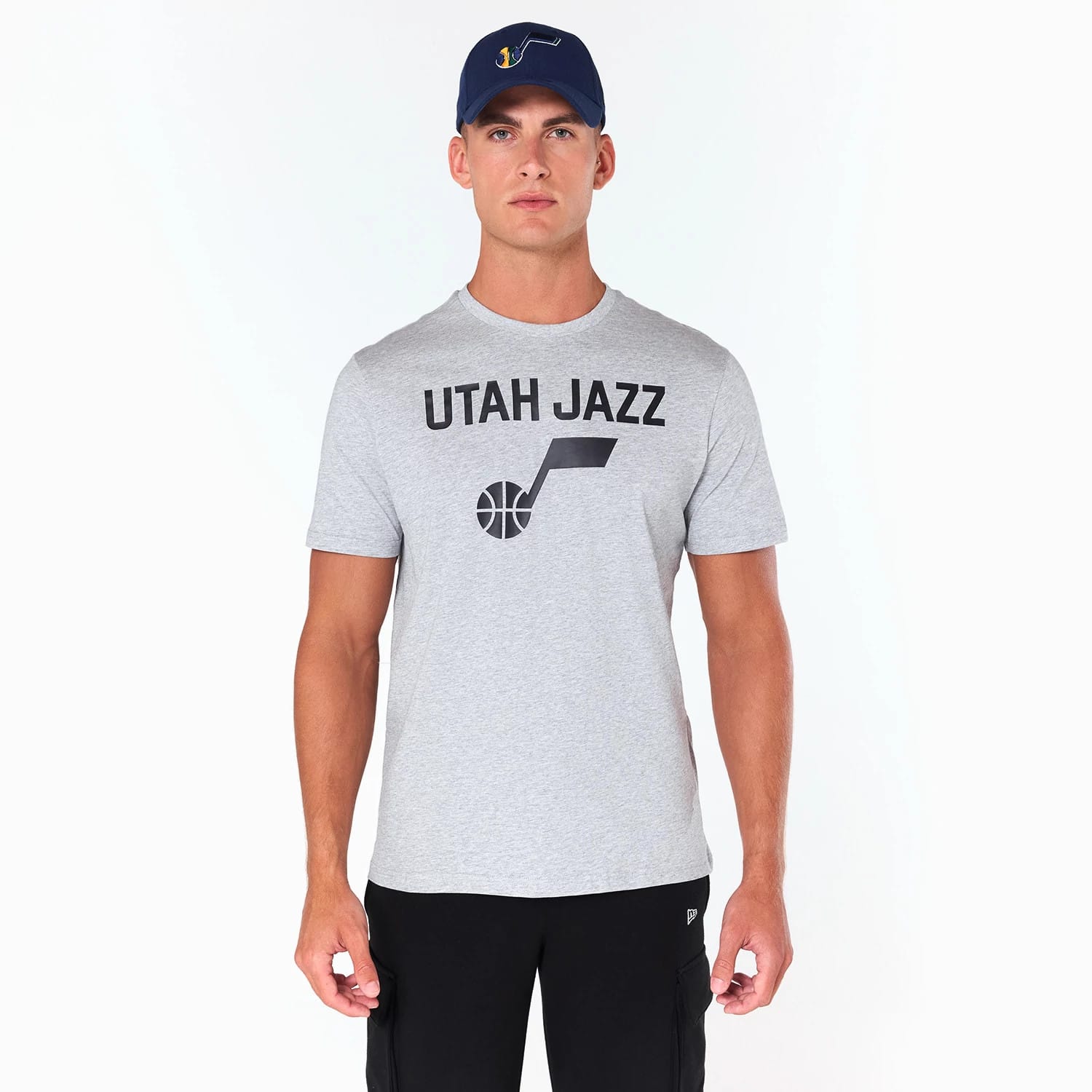 New Era Utah Jazz NBA Grey T-Shirt, on-model view showing the grey T-shirt with black 'Utah Jazz' text and logo printed on the chest, complemented by a raised logo on a dark blue cap worn by the model.