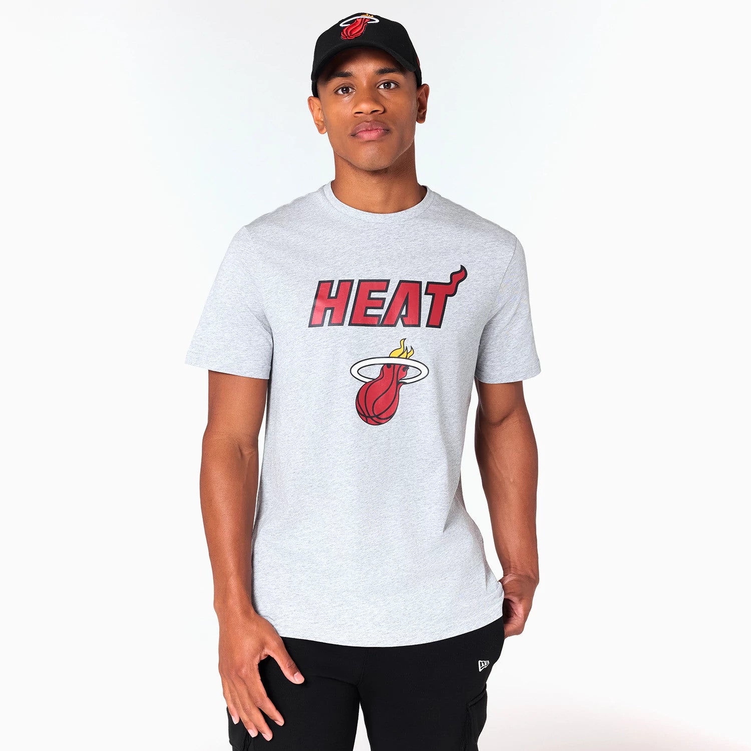 New Era Miami Heat NBA Grey T-Shirt, on-model view showing the grey fabric with a large red and black Heat logo on the chest, the model wearing a black cap featuring an embroidered team emblem, paired with black trousers.