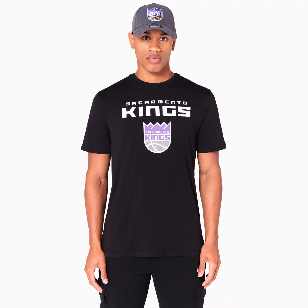 New Era Sacramento Kings NBA Black T-Shirt, front view showing the black fabric with the Sacramento Kings logo printed in purple and white across the chest and a model also wearing a grey cap with a raised Kings patch on the front.
