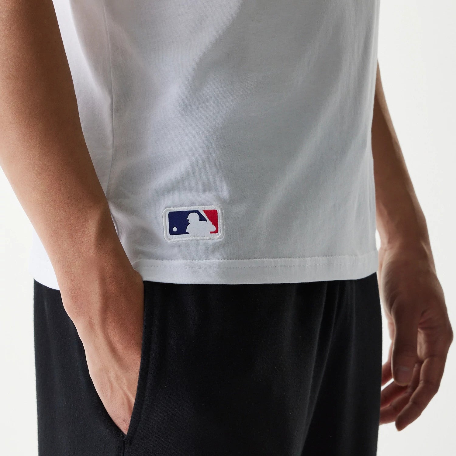 New Era MLB Generic Logo MLB Regular White T-Shirt, side view showing the lower half of a white T-shirt with a raised MLB logo patch above the hem on the left side near the seam, paired with black trousers.