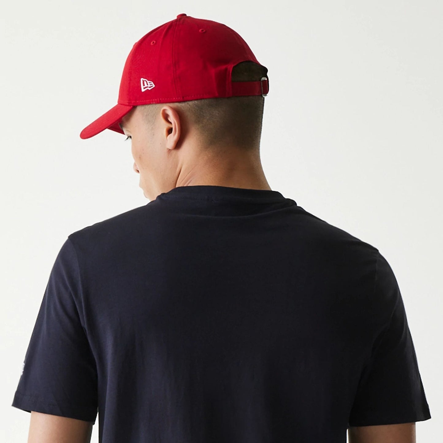 New Era Cleveland Guardians MLB Regular Navy T-Shirt, on-model view showing the plain navy back of the T-shirt with a red cap on the model's head, featuring a raised New Era logo on the side of the cap.
