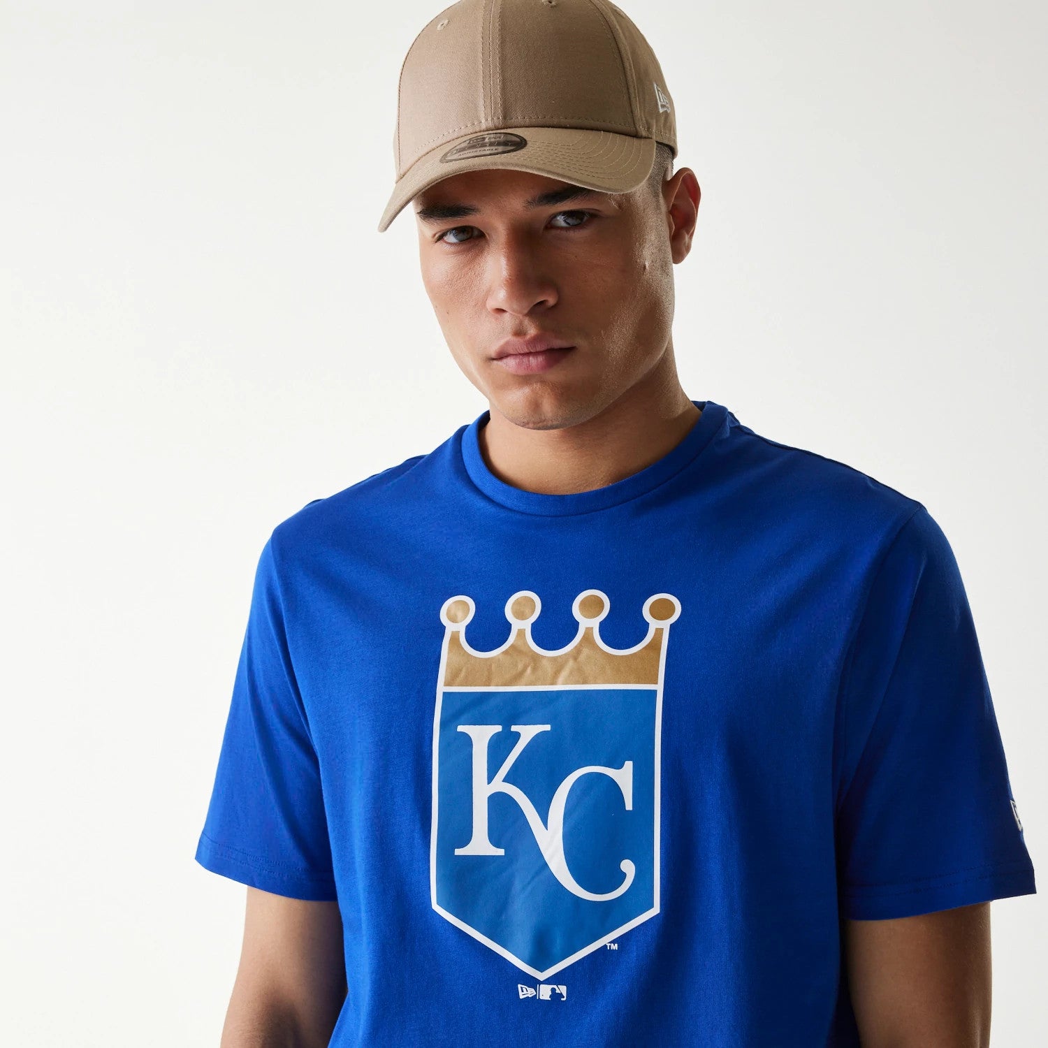New Era Kansas City Royals MLB Regular Blue T-Shirt, front view showing a large KC and crown emblem in white and gold on the chest, worn by a model in a beige cap with a debossed logo on the side.