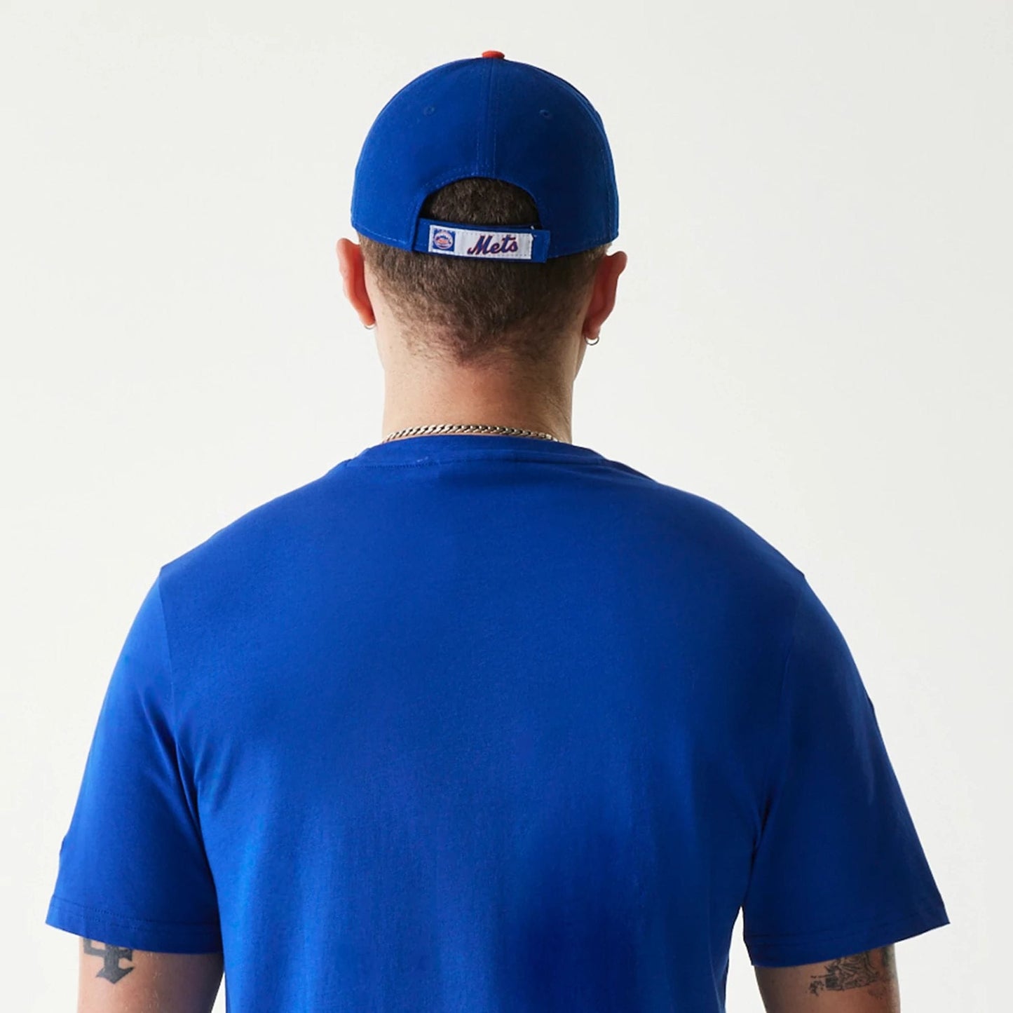 New Era New York Mets MLB Regular Blue T-Shirt, on-model rear view showing the solid blue fabric covering the back, a Mets logo on a blue cap worn backwards with eyelets on the crown above the band.