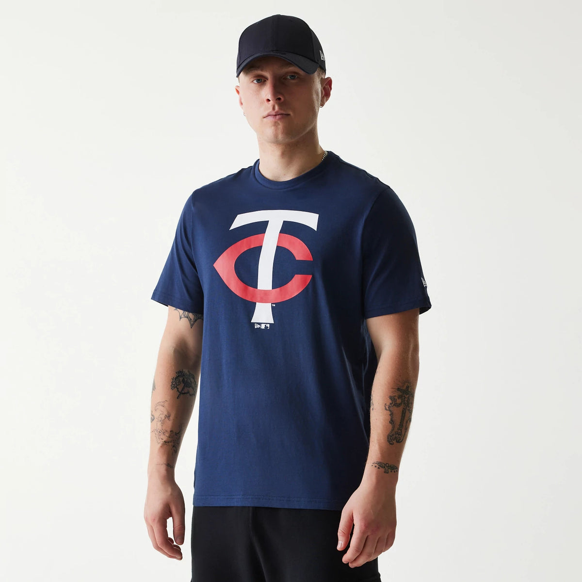 New Era Minnesota Twins MLB Regular Dark Blue T-Shirt, on-model view showing a large white and red logo printed on the chest, worn by a person in black trousers and a plain black cap, with the New Era logo embroidered on the cap's side.