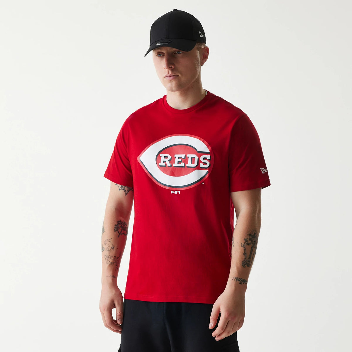 New Era Cincinnati Reds MLB Regular Red T-Shirt, on-model view showing a large central white and red Reds logo on the chest, a New Era logo embroidered in white on the sleeve, worn with a plain black cap.